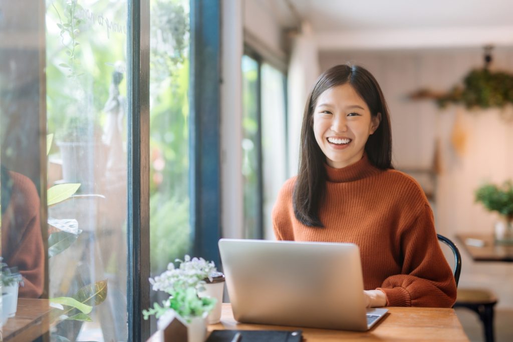 Woman smiling while working on a laptop in a bright, plant-filled café.