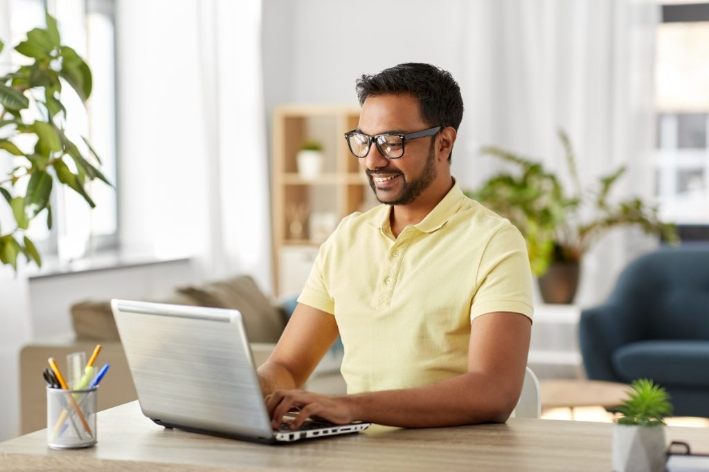 Man wearing glasses working on a laptop at a desk in a bright home office.
