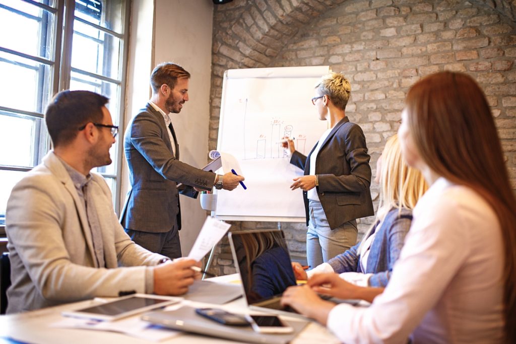 Team gathered in a meeting room while two presenters stand at a flip chart reviewing a hand-drawn bar graph, with others watching and taking notes.