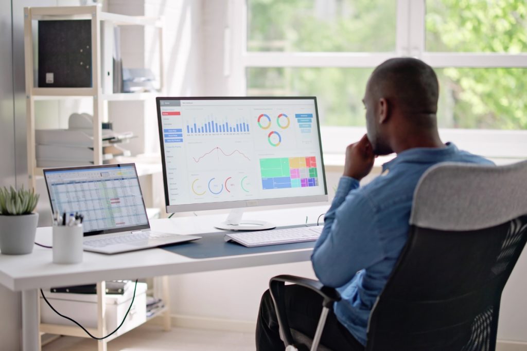 Man sitting at a desk reviewing charts and dashboards on a large computer monitor, with a laptop and office supplies beside him.